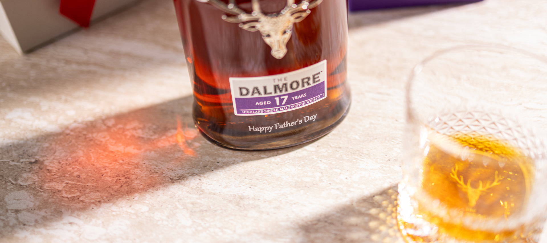 Bottle of The Dalmore 17 Year Old single malt Scotch whisky with stag emblem and “Happy Father’s Day” message, next to a whisky glass and presentation box on a stone surface.