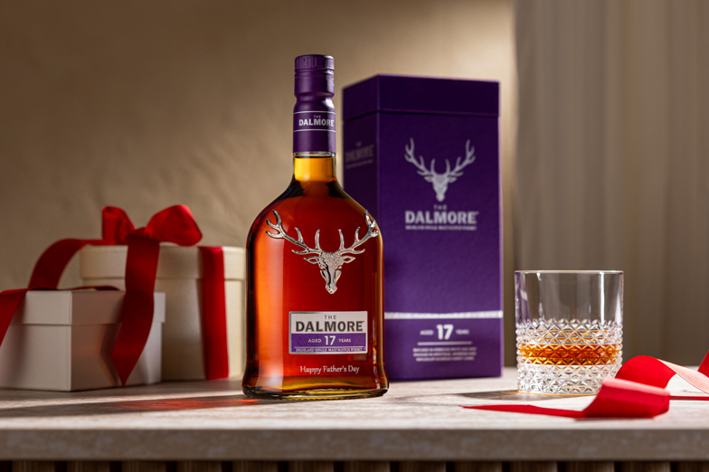 The Dalmore 17 Year Old single malt Scotch whisky bottle with stag emblem, gift boxes with red ribbons, a branded presentation box, and a whisky glass on a wooden surface.